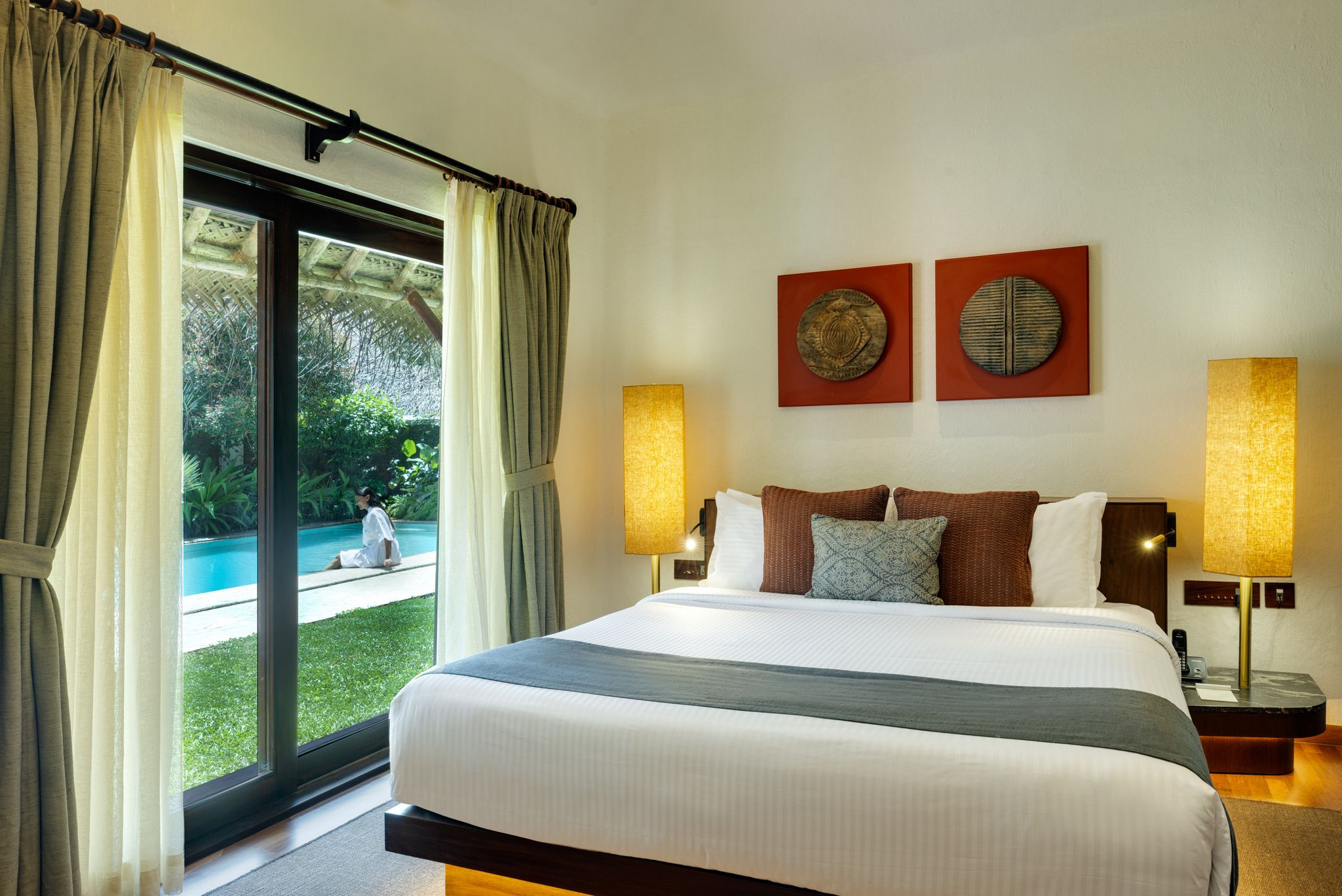 Quarto de hotel com uma cama grande, duas luminárias na parede, vista para uma piscina e uma porta de correr de vidro.