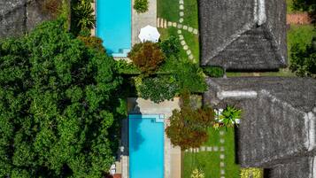 Vista aérea de uma propriedade com duas piscinas, rodeada por vegetação exuberante e construções com telhados de palha.