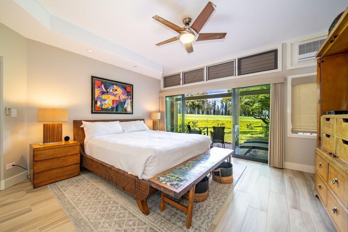 Ein Schlafzimmer mit einem Bett, Nachttischen, einer Bank, einem Deckenventilator, einem Gemälde und Blick auf einen Garten durch große Fenster.