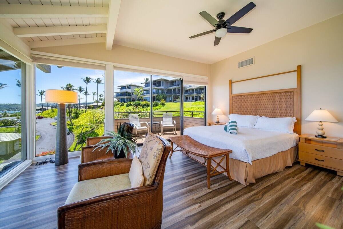 Ein Schlafzimmer mit einem großen Bett, einem Deckenventilator, Blick auf ein Gebäude und Grünfläche sowie einem Balkon mit Stühlen und einem Tisch.