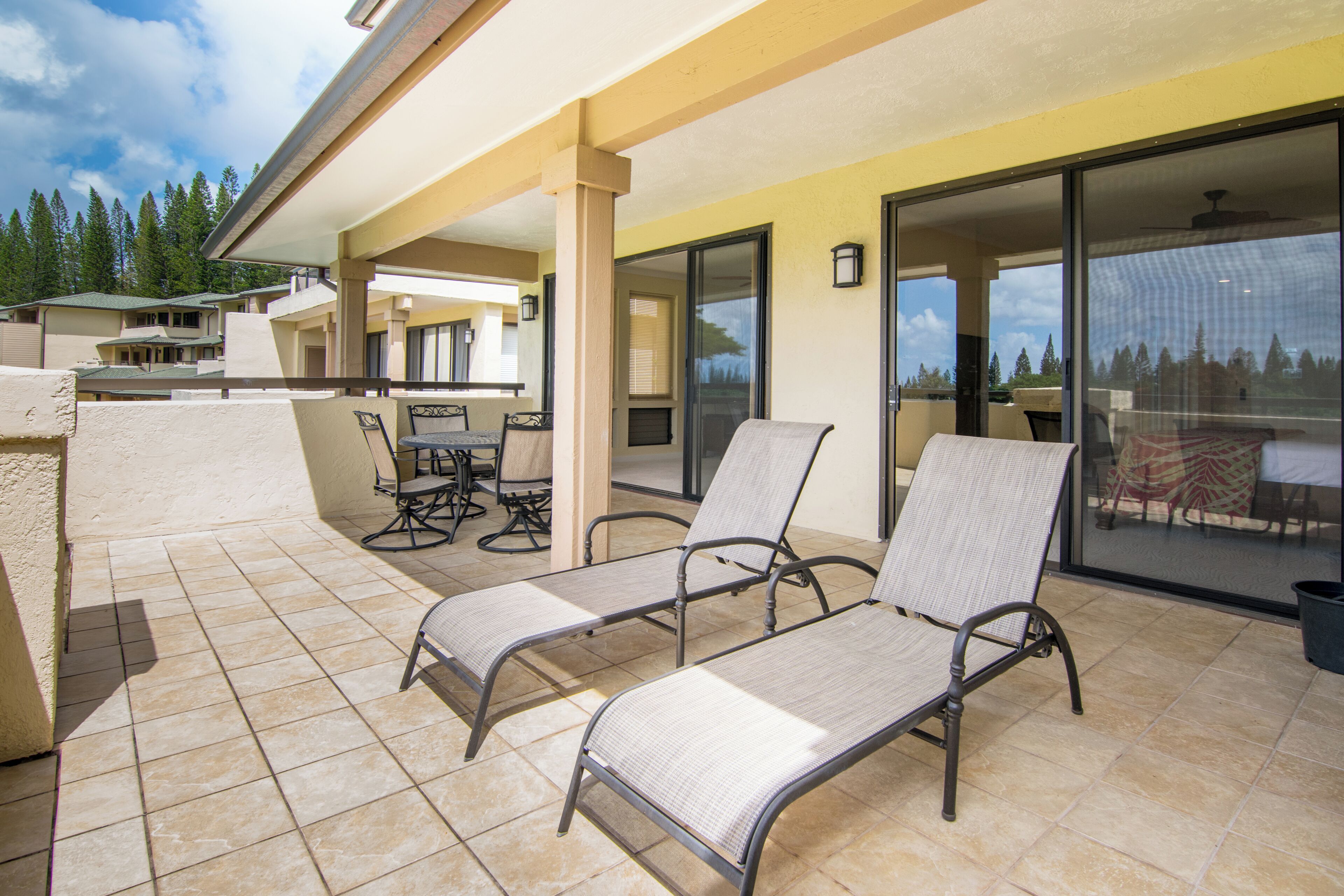 A covered patio area with two lounge chairs, a table, and chairs, and a sliding glass door leading to a balcony with a view of trees.