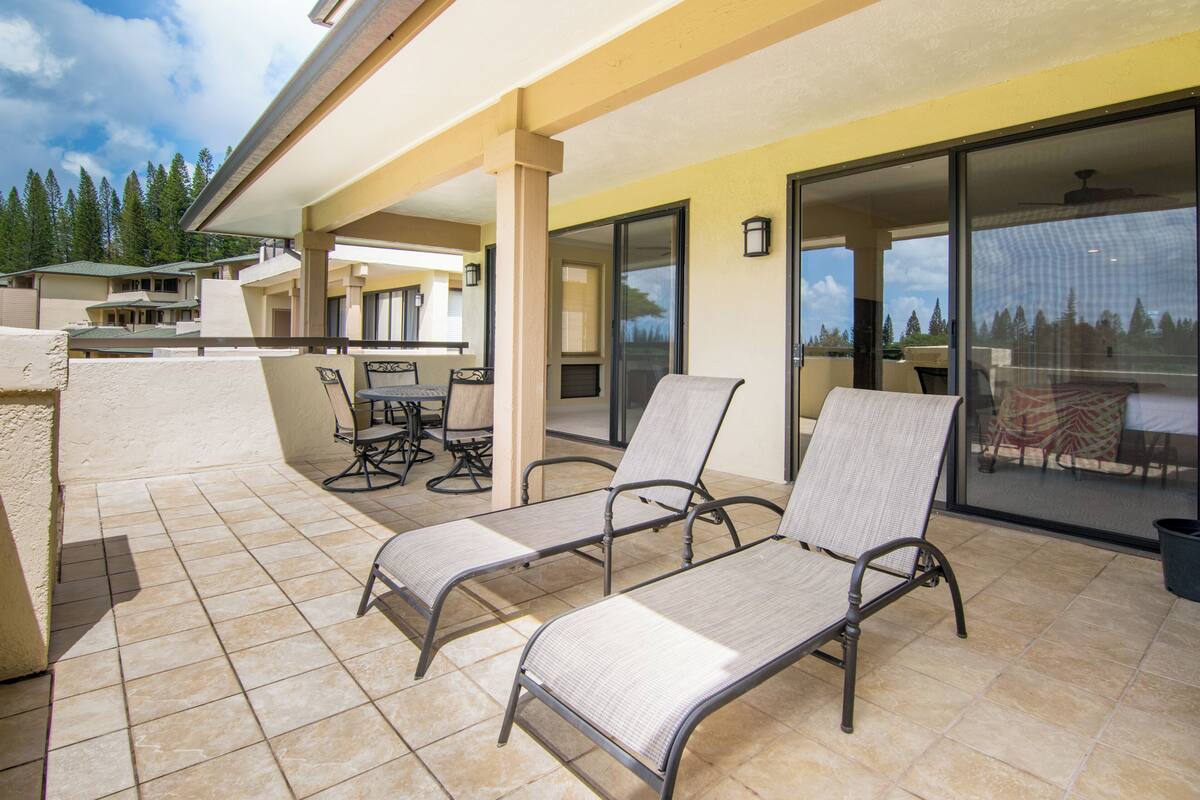 Ein überdachter Patio-Bereich mit zwei Liegestühlen, einem Tisch und Stühlen, sowie eine Glasschiebetür, die auf einen Balkon mit Blick auf Bäume führt.