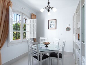 A dining area with a glass table, white chairs, a chandelier, and a view through a window with open shutters. - Los Pinos 25 Villas by Casasol by Interhome (Nerja)