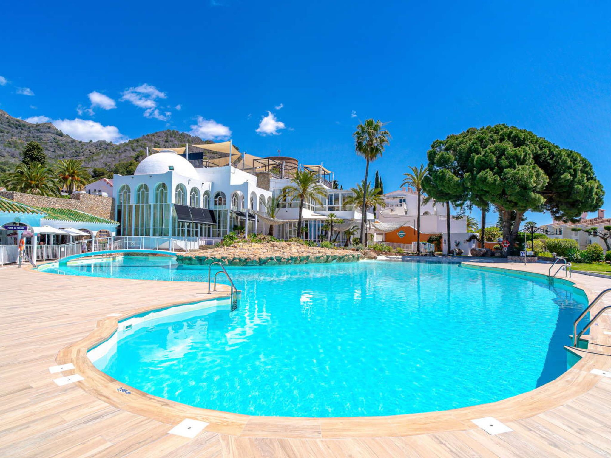 Una gran piscina exterior con terraza de madera, rodeada de palmeras y un edificio blanco con ventanas de arco.