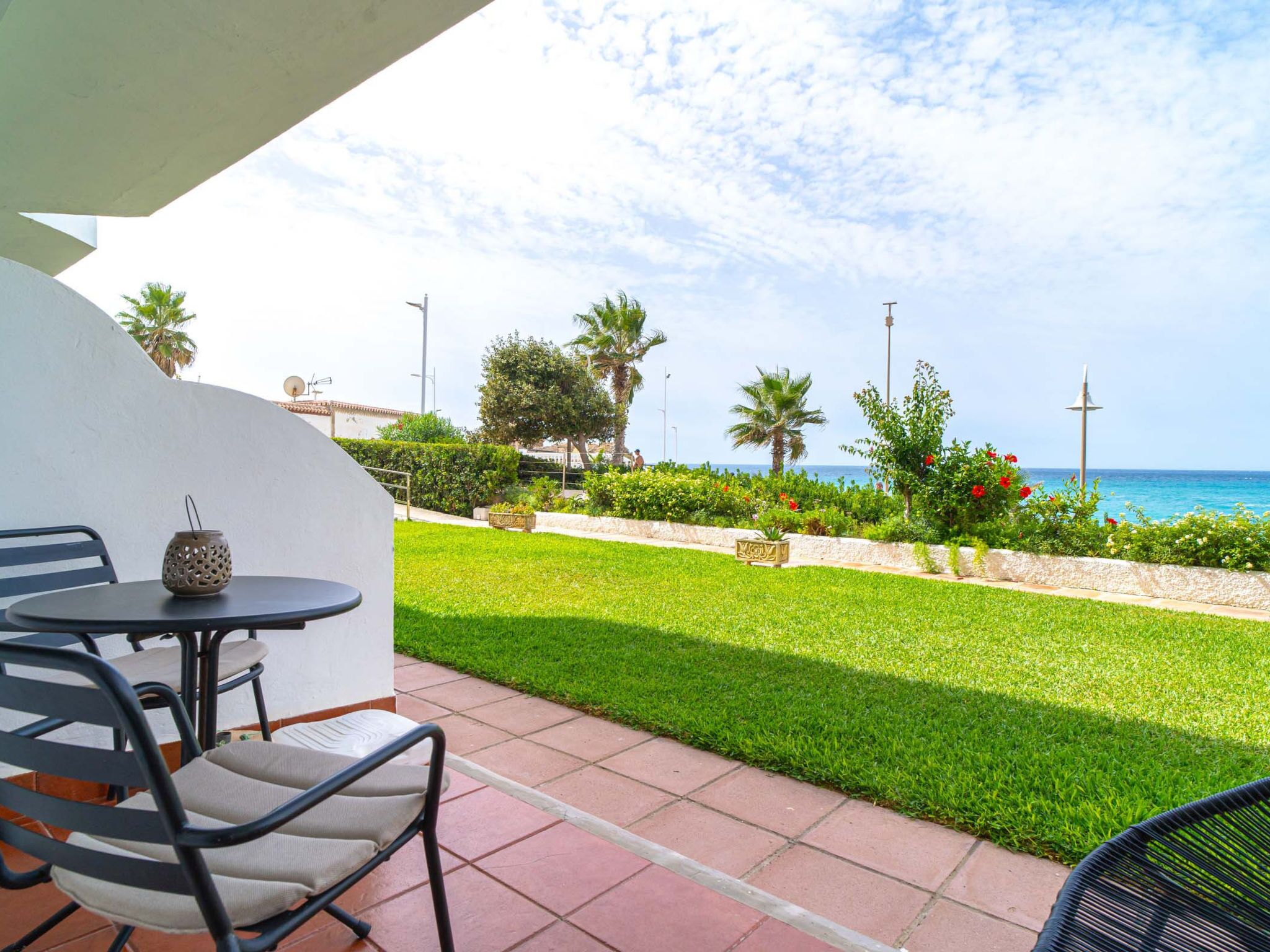 A patio with outdoor seating, a view of the sea, and a well-maintained garden with palm trees and red flowers.