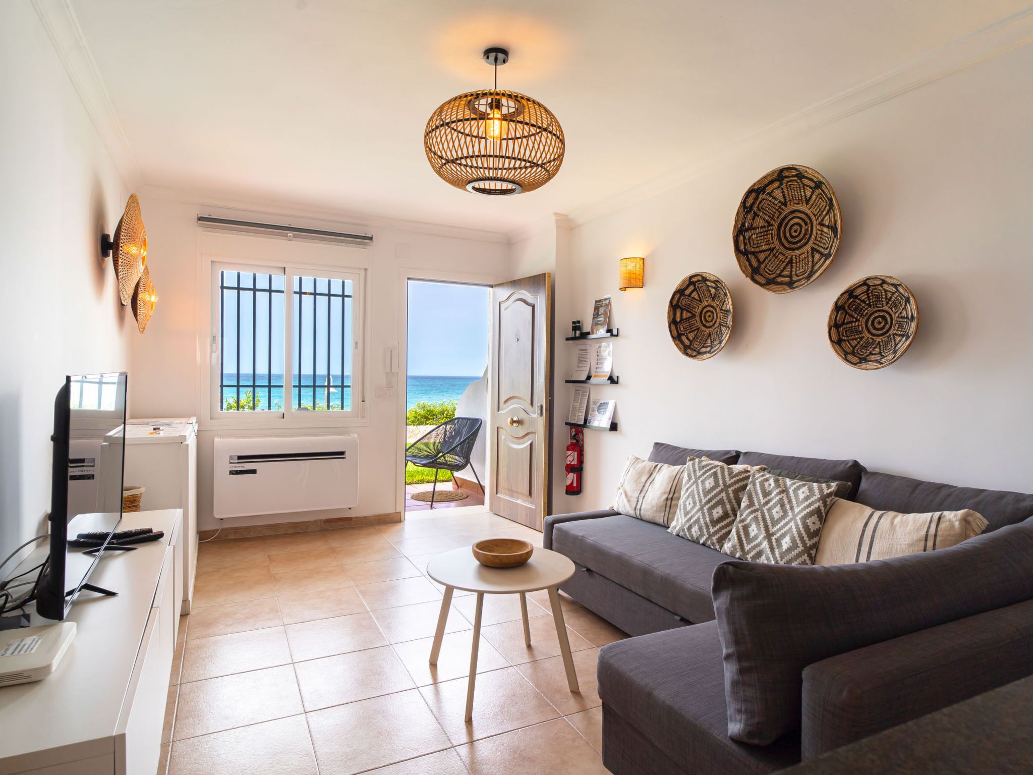 Een woonkamer met een grijze bank, een kleine ronde tafel en een televisie op een witte kast. Er hangen decoratieve borden aan de muur en een raam met uitzicht op zee.