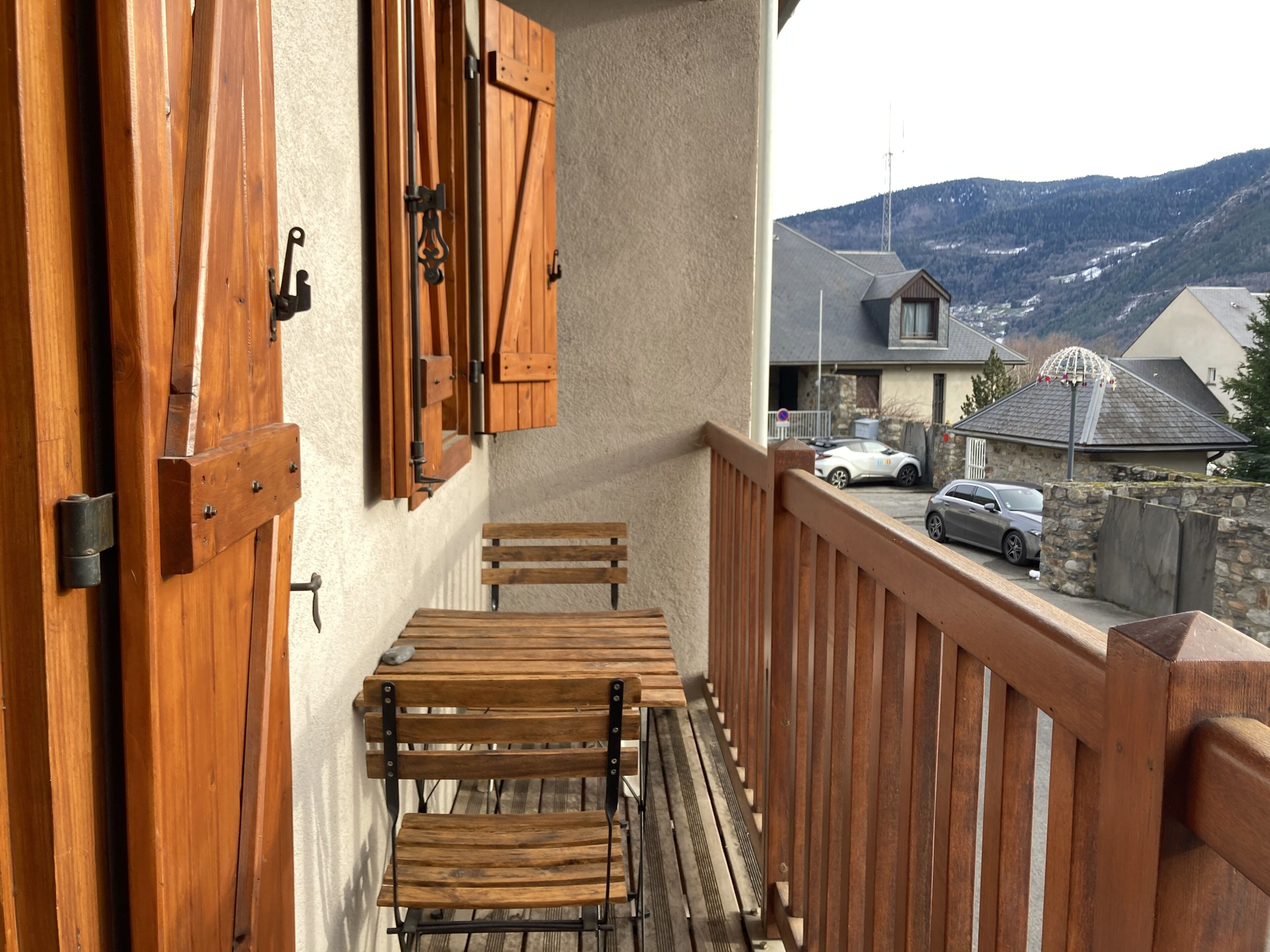 A wooden balcony with a table and chairs, a view of a mountain, and parked cars.