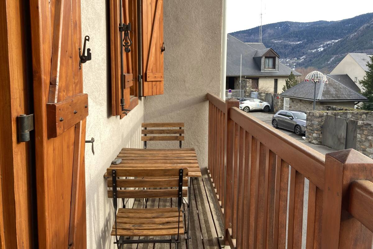 Un balcón de madera con una mesa y sillas, vistas a una montaña y coches aparcados.
