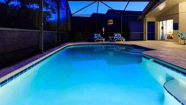 A swimming pool with a covered area, two lounge chairs, and a building in the background. - Disney Area Budget Homes (Davenport)