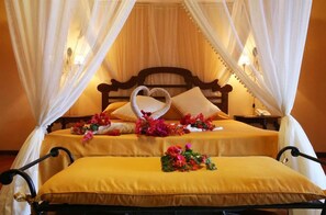 A double bed with a heart-shaped pillow arrangement, a bedside table with a lamp, and a canopy bed with draped curtains. - Karafuu Hotel Beach Resort (Michamvi)