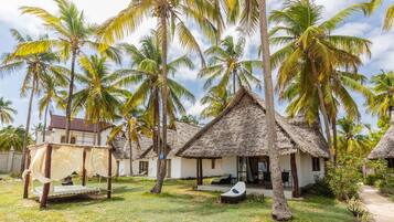 A thatched-roof bungalow with a small porch, surrounded by palm trees and a grassy lawn.