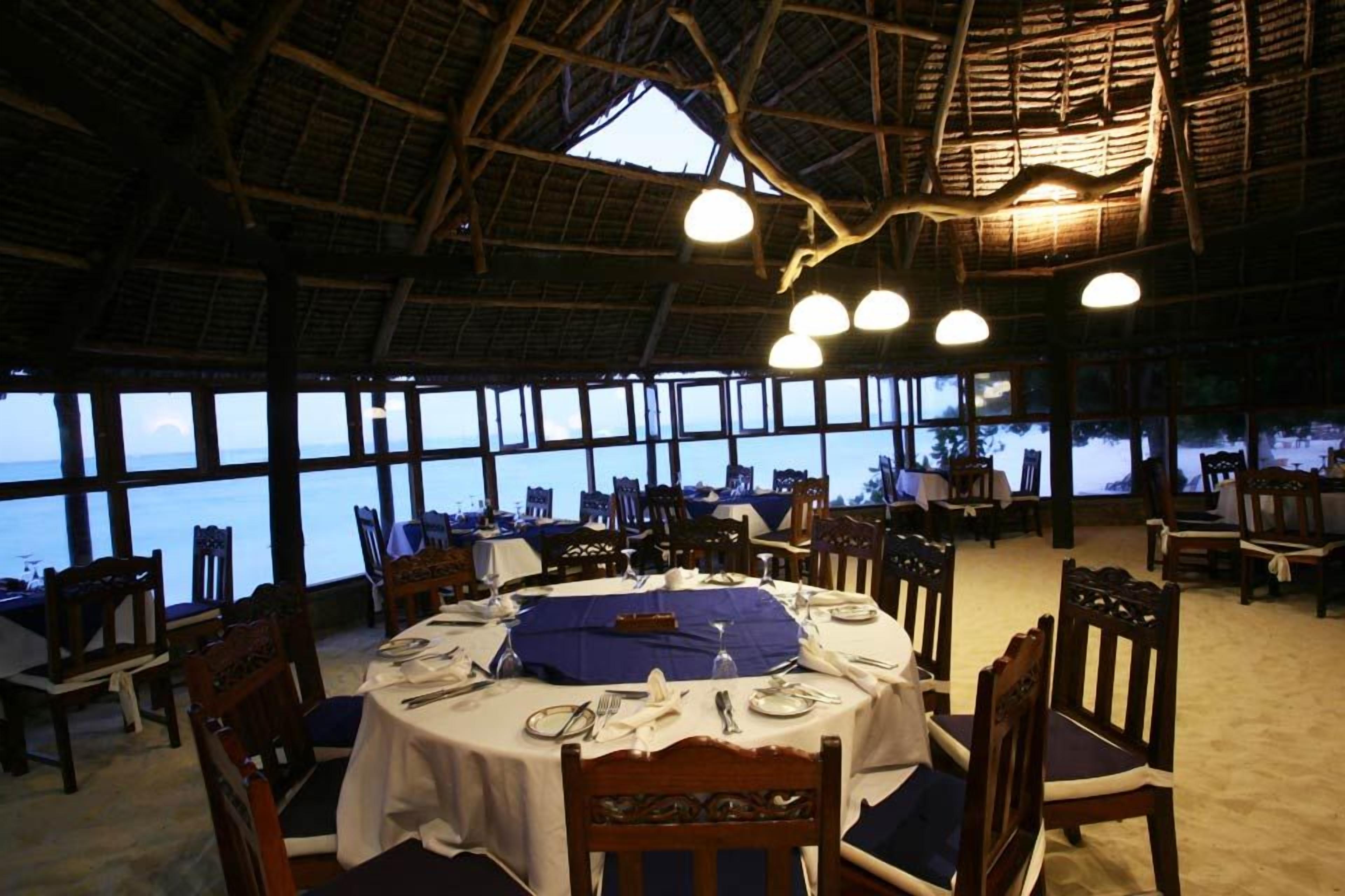 Restaurante com mesas redondas preparadas para refeições, grandes janelas com vista para o mar e estrutura de telhado de palha.