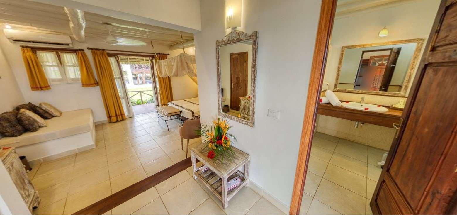 Habitación de hotel con cama, zona de estar con sofá, una mesita con un florero, un espejo y un baño con lavamanos y espejo.