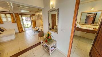 Habitación de hotel con cama, zona de estar con sofá, una mesita con un florero, un espejo y un baño con lavamanos y espejo.