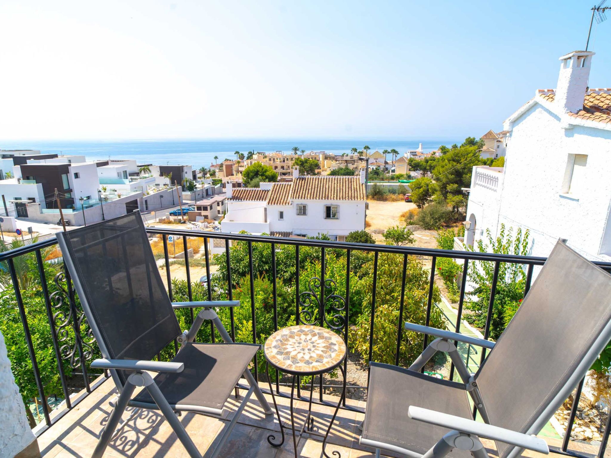 Un balcon avec deux chaises et une petite table, donnant sur une ville côtière avec ses bâtiments et offrant une vue sur la mer.