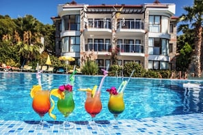 A pool with four colorful drinks, a building with balconies, and palm trees in the background. - Tropical Deluxe Suites (Fethiye)