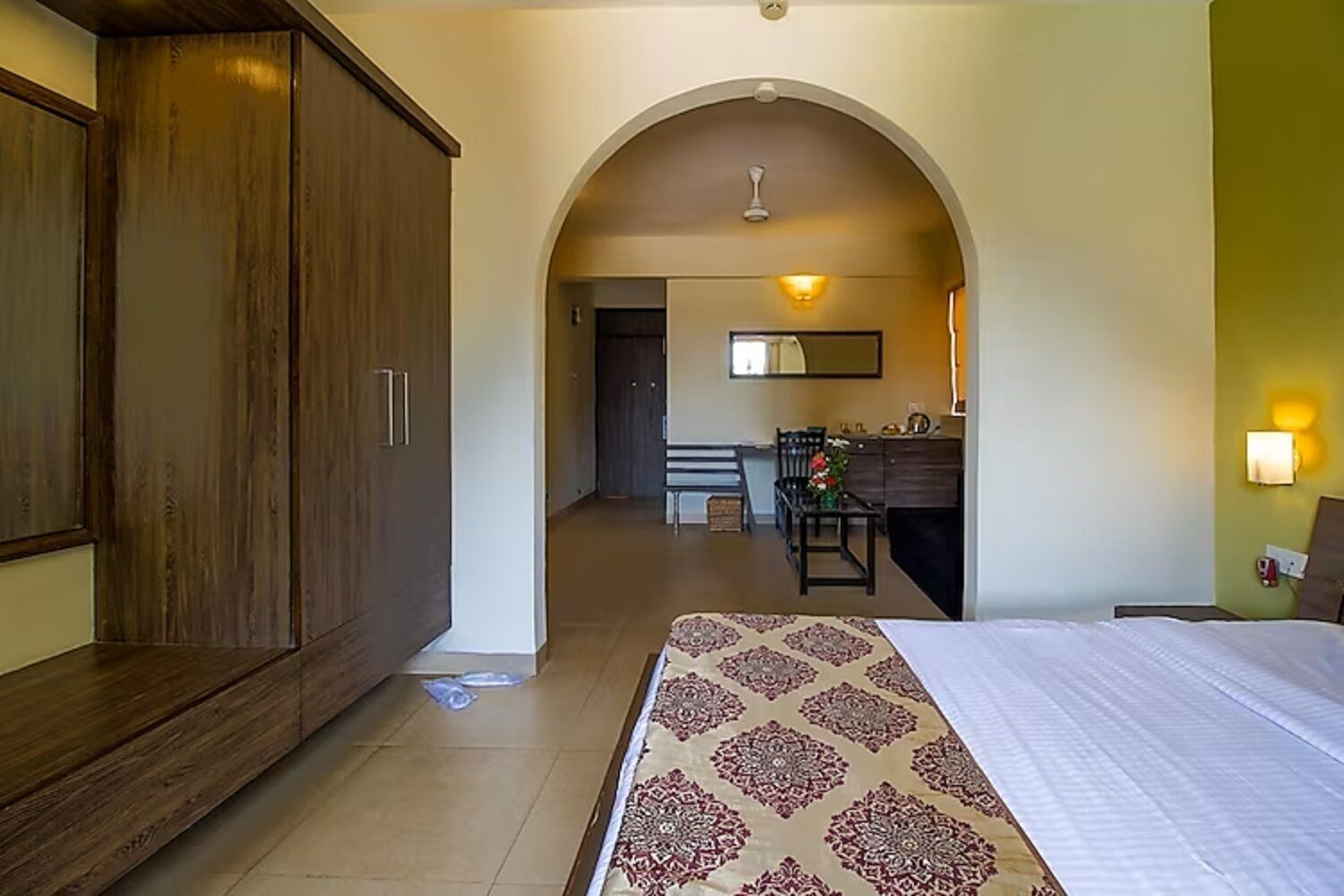 A hotel room with a bed, a wardrobe, a dining area with a table and chairs, and an arched doorway leading to another room.