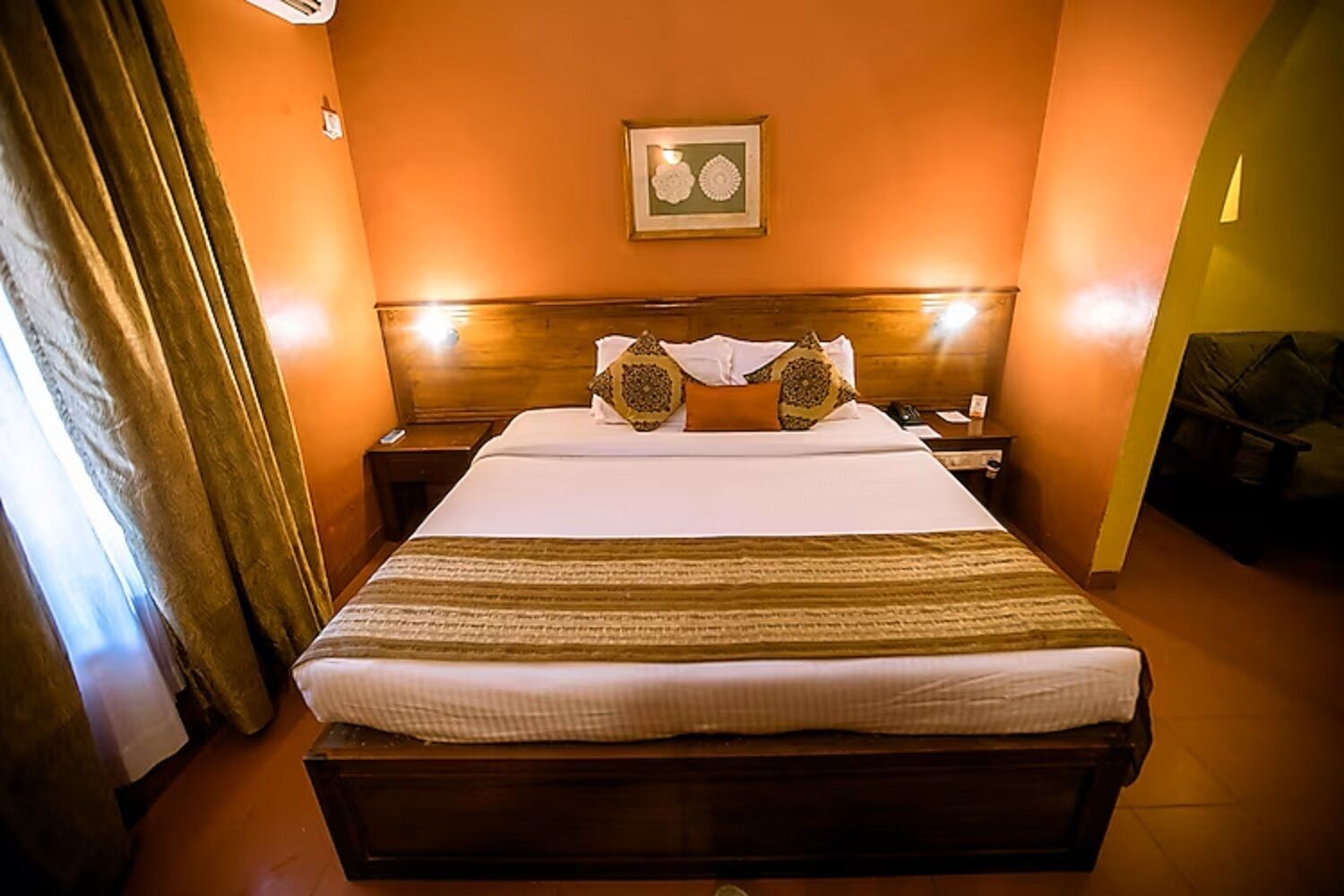A hotel room with a large bed, a wooden headboard, a framed picture on the wall, and a curtain on the left side.