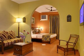A room with a sofa, a wooden chest of drawers, a chair, a bed with a patterned bedspread, a lamp, and a television. - Pride Sun Village Resort (Arpora)