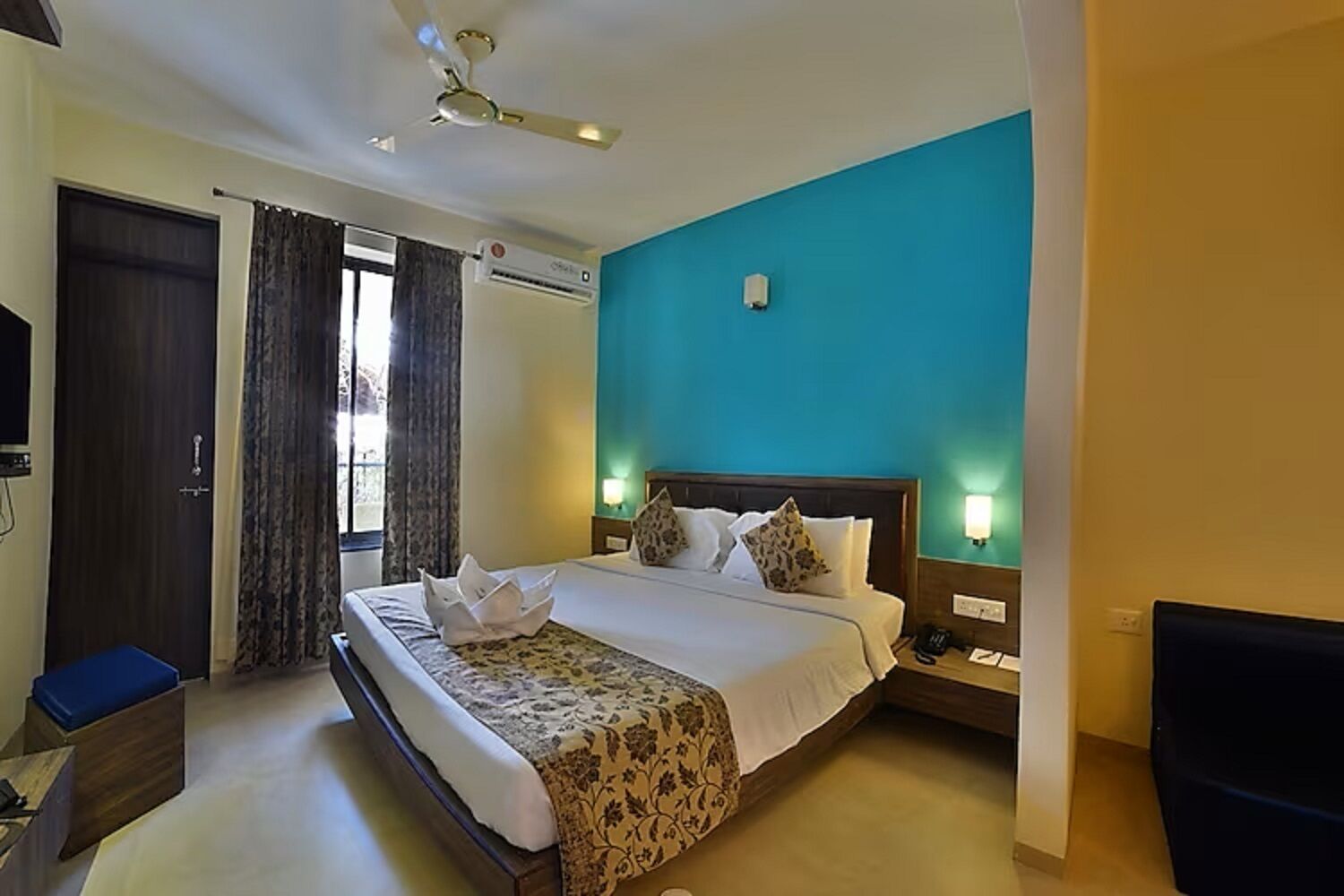 A hotel room with a bed, a desk, a chair, a ceiling fan, and a window with curtains.