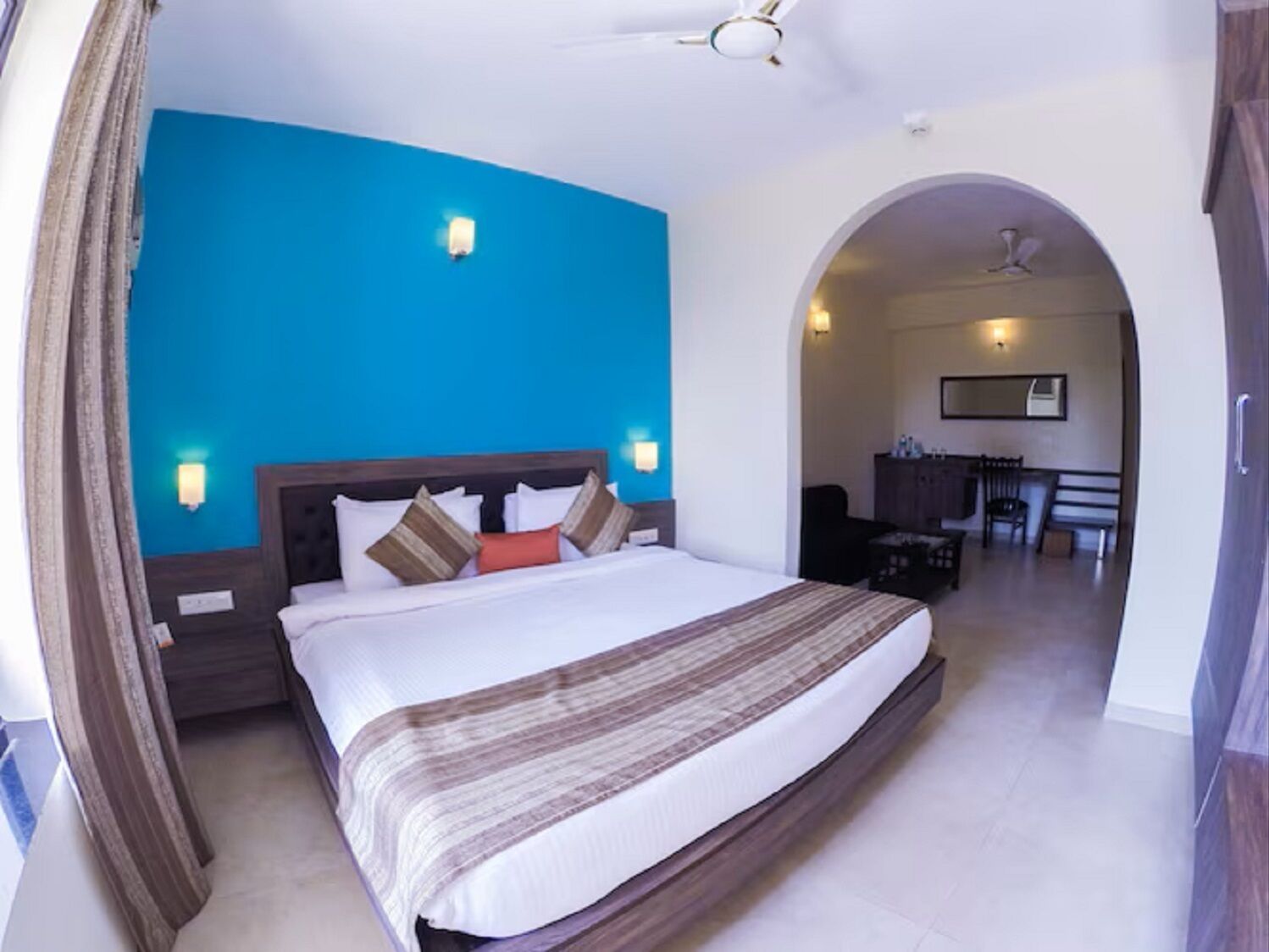 A hotel room with a large bed, a ceiling fan, and a view of the dining area through an archway.