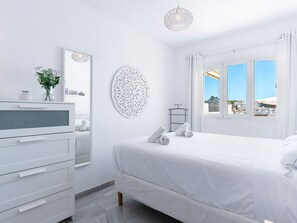 A white bedroom with a bed, a dresser, a mirror, a decorative wall piece, and a window with a view. - Apartment  by Interhome (Nerja)