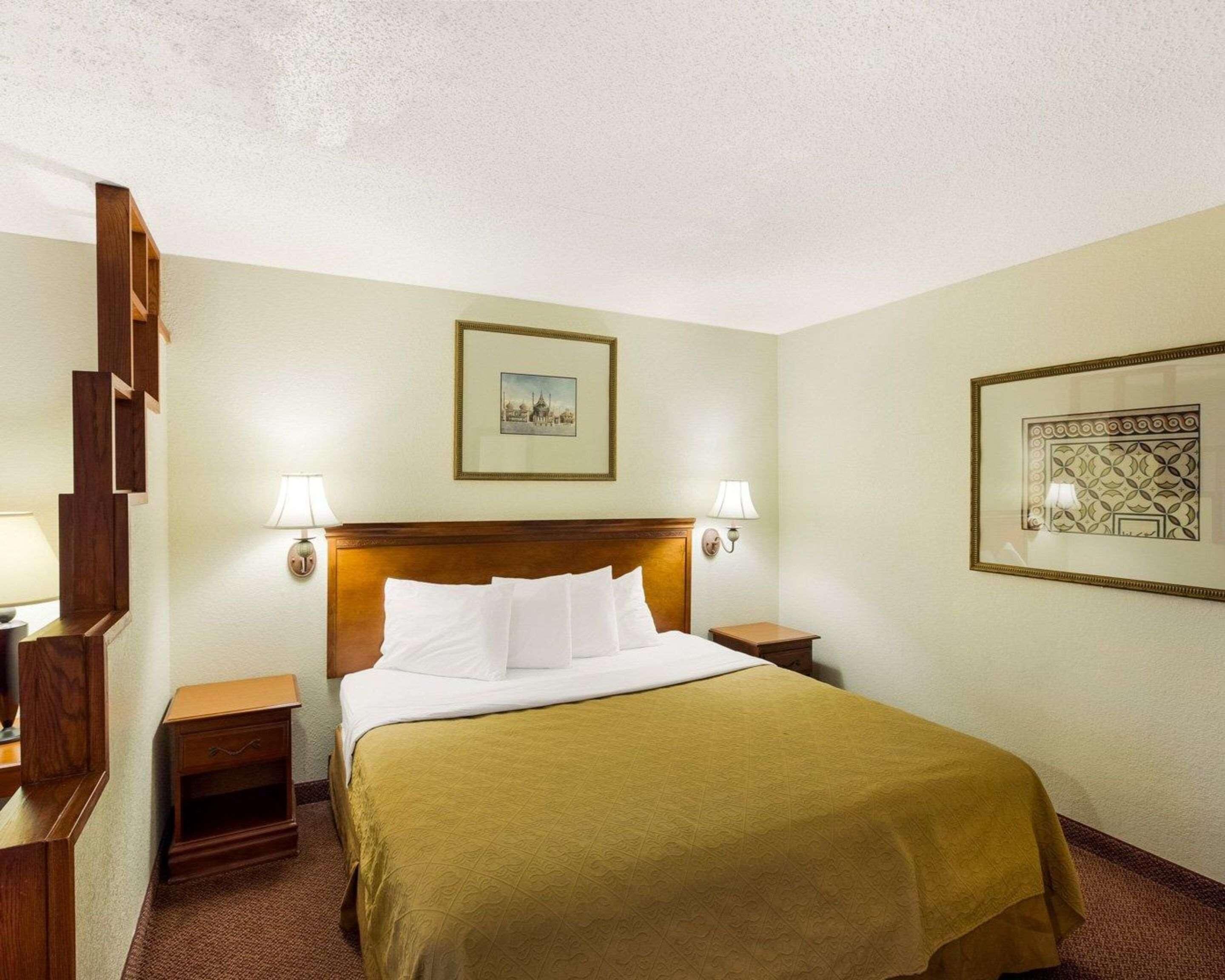 A hotel room with a bed, two bedside tables, a headboard, wall-mounted lamps, and framed pictures.