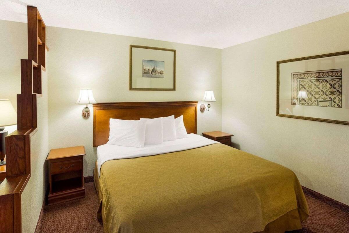 A hotel room with a bed, two bedside tables, a headboard, wall-mounted lamps, and framed pictures.