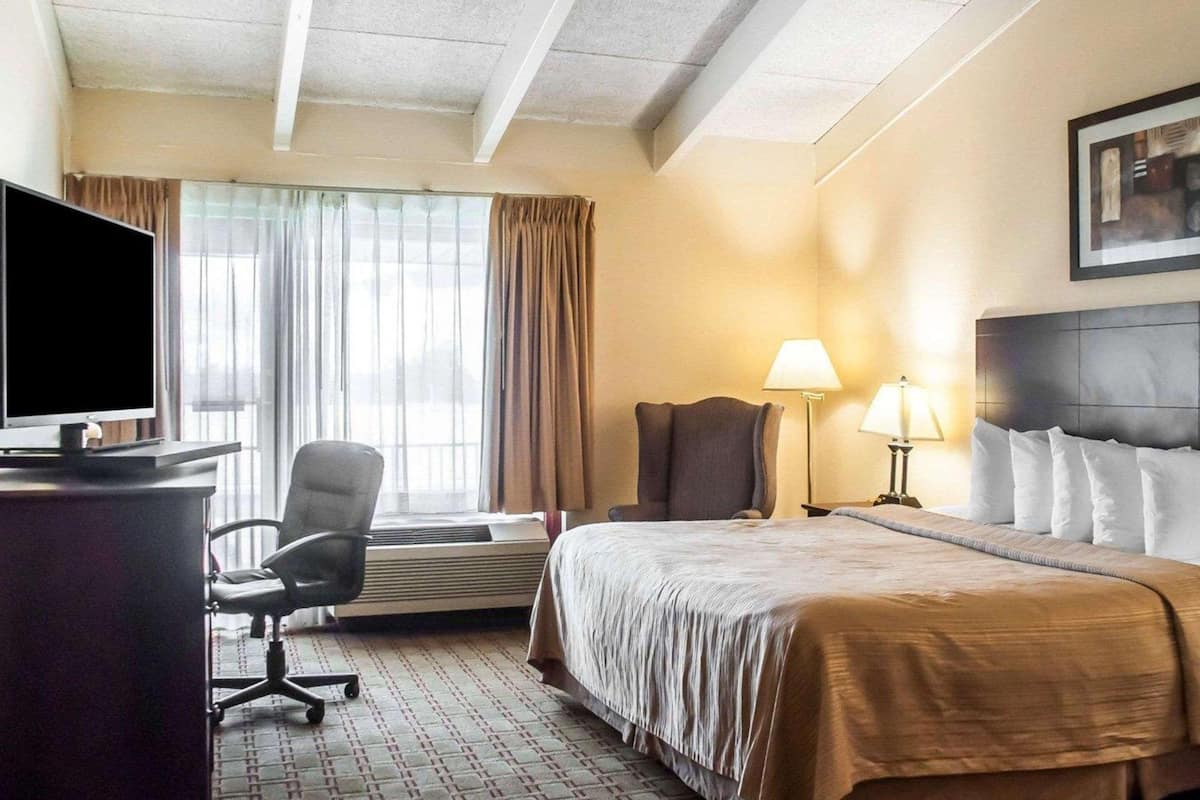 A hotel room with a bed, a desk with a TV, a chair, a lamp, and a window with curtains.