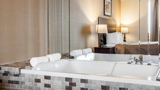 Un baño de hotel con una bañera grande, un espejo, una mesita de noche con lámpara y una cama con cabecero.