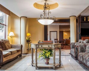 Un hall d’hôtel avec un lustre classique, un canapé, une table ornée d’un vase de fleurs et un téléviseur en arrière-plan.