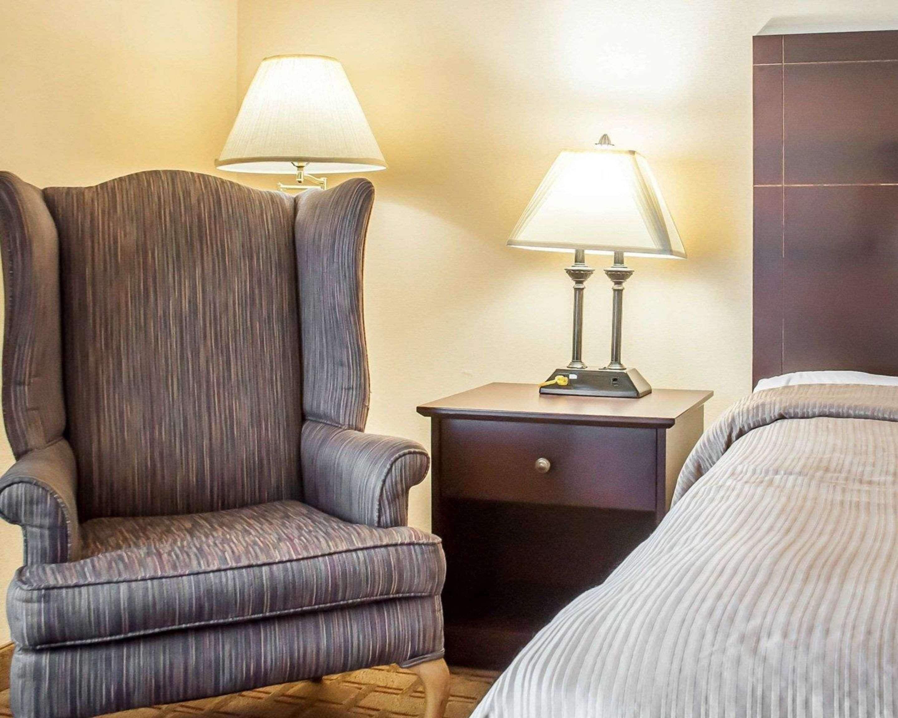 Habitación de hotel con un sillón grande, una mesita de noche con lámpara y una cama con colcha a rayas.