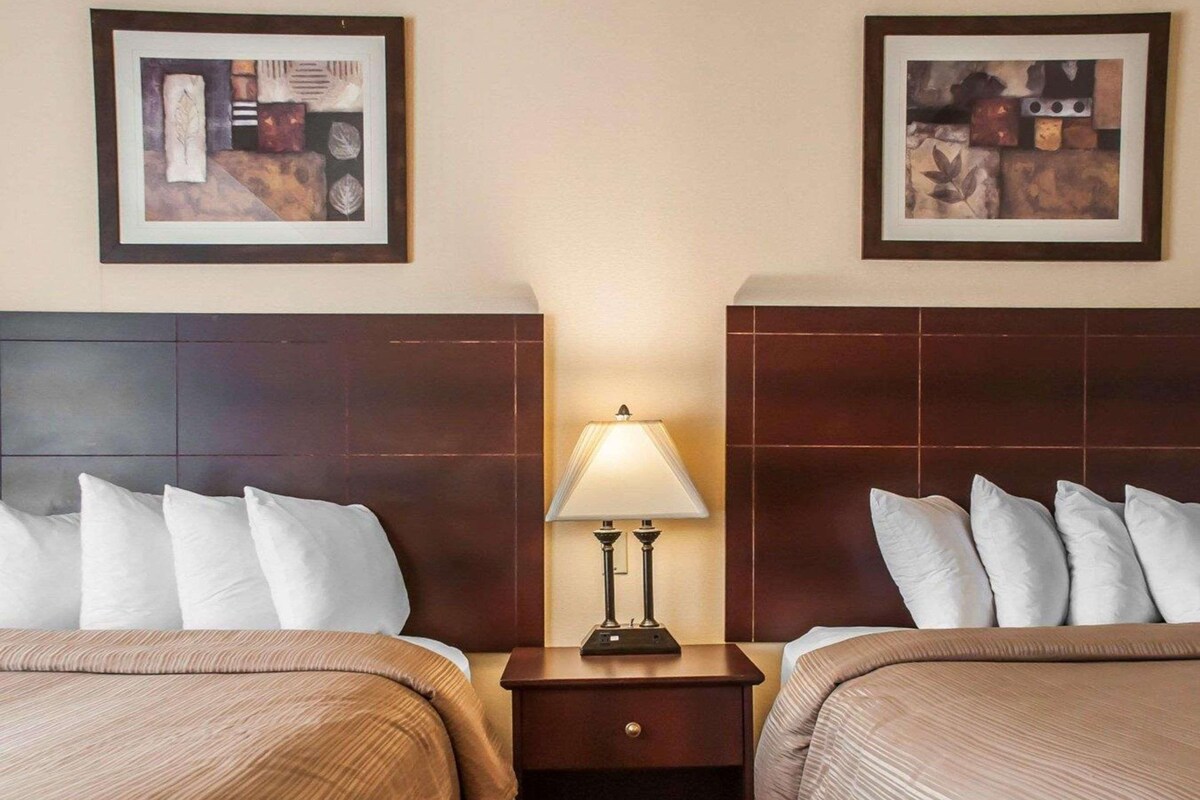 A hotel room with two beds, a nightstand with a lamp, and two framed artworks on the walls.