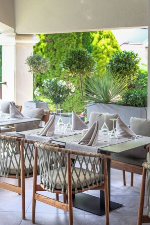 An outdoor dining area with wicker chairs and a table set for two, featuring a view of lush greenery and a modern building in the background. - Grifid Hotel Bolero (Varna)
