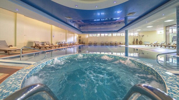 Una piscina interior con borde de azulejos, sillas de descanso e iluminación empotrada en el techo.