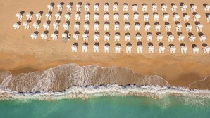 An aerial view of a beach with rows of white umbrellas and beach chairs, a sandy shoreline, and waves approaching the shore. - Grifid Hotel Bolero (Varna)