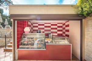 Un quiosco de comida con pared de baldosas blancas y rojas en damero, un expositor de vidrio con pasteles y una pequeña barra con una máquina de café.