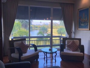 A room with two cushioned chairs, a small round table, and a large window offering a view of a golf course and water. - Mission Hills Phuket Golf Resort & Spa (Wichit)