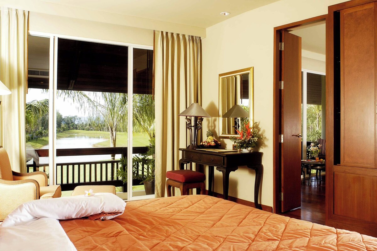 A hotel room with a bed, a desk, a chair, a mirror, and a view of a garden.