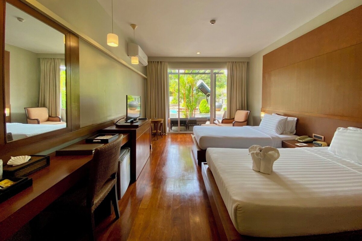 A hotel room with a wooden desk, two beds, a large mirror, and a view of a pool and greenery.