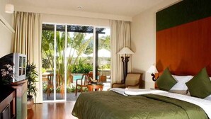 A hotel room with a bed, a desk, a chair, a lamp, and a view of a pool and palm trees.