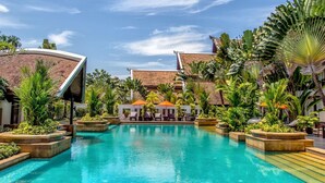 A pool area with lounge chairs and umbrellas, surrounded by tropical plants and buildings with curved roofs. - Mission Hills Phuket Golf Resort & Spa (Wichit)