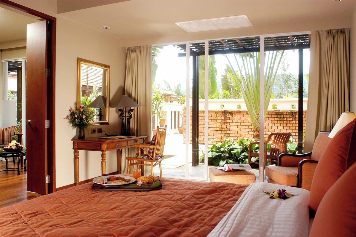 A hotel room with a bed, a desk, a chair, and a balcony with plants.