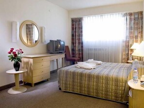 Habitación de hotel con cama, escritorio con televisor, espejo, florero con flores y ventana con cortinas.