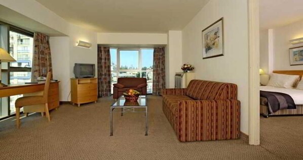 Habitación de hotel con sofá, una mesita con un florero, televisor, escritorio y ventana con vista a edificios.