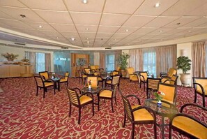 Un salón de conferencias de hotel con alfombra roja estampada, varias mesas y sillas, y ventanas grandes con cortinas.