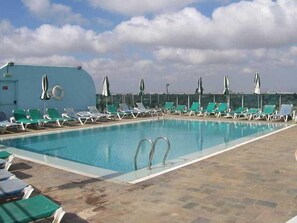 Una piscina con sillas de descanso y sombrillas, un edificio azul y un cielo despejado.