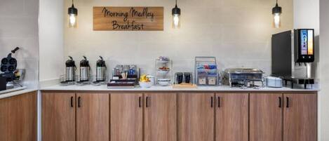 A breakfast area with a sign reading 'Morning Mellow Breakfast,' equipped with coffee machines, a fruit bowl, and a water dispenser.