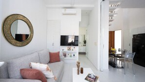 En moderne stue med en grå sofa, et rundt spejl, et fladskærms-tv og et spiseområde med et glasbord og stole.