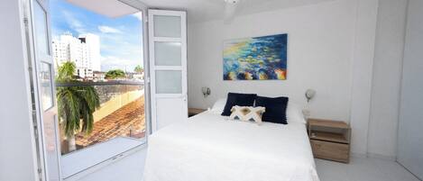 A bedroom with a bed, a nightstand, a painting, and a view of buildings through a sliding door.